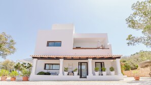 Une maison moderne à deux étages, avec une façade blanche, un balcon et des plantes en pot disposées autour de l’entrée.
