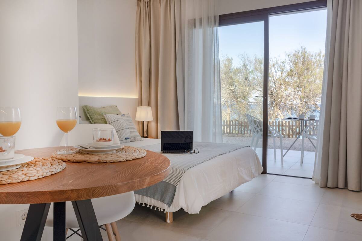 Un dormitorio con cama, una computadora portátil, una mesa con vasos de jugo de naranja y vistas al exterior a través de una puerta corredera.