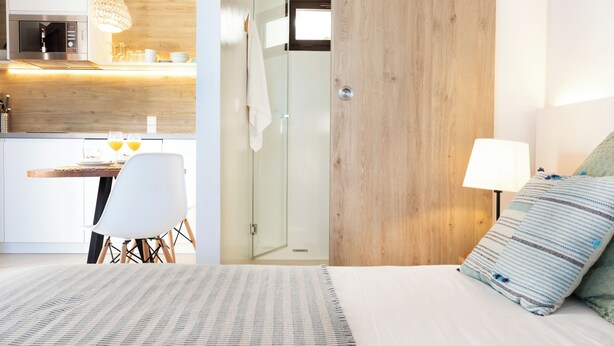Une chambre d’hôtel moderne avec un mur d’accent en bois, un lit avec un oreiller à rayures, une lampe de chevet et une vue sur la cuisine, où l’on aperçoit un four à micro-ondes et un luminaire suspendu.