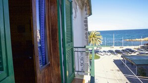 Un edificio costero con puerta de madera y contraventanas verdes, un balcón con barandilla de metal, y un estacionamiento con coches y una palmera.