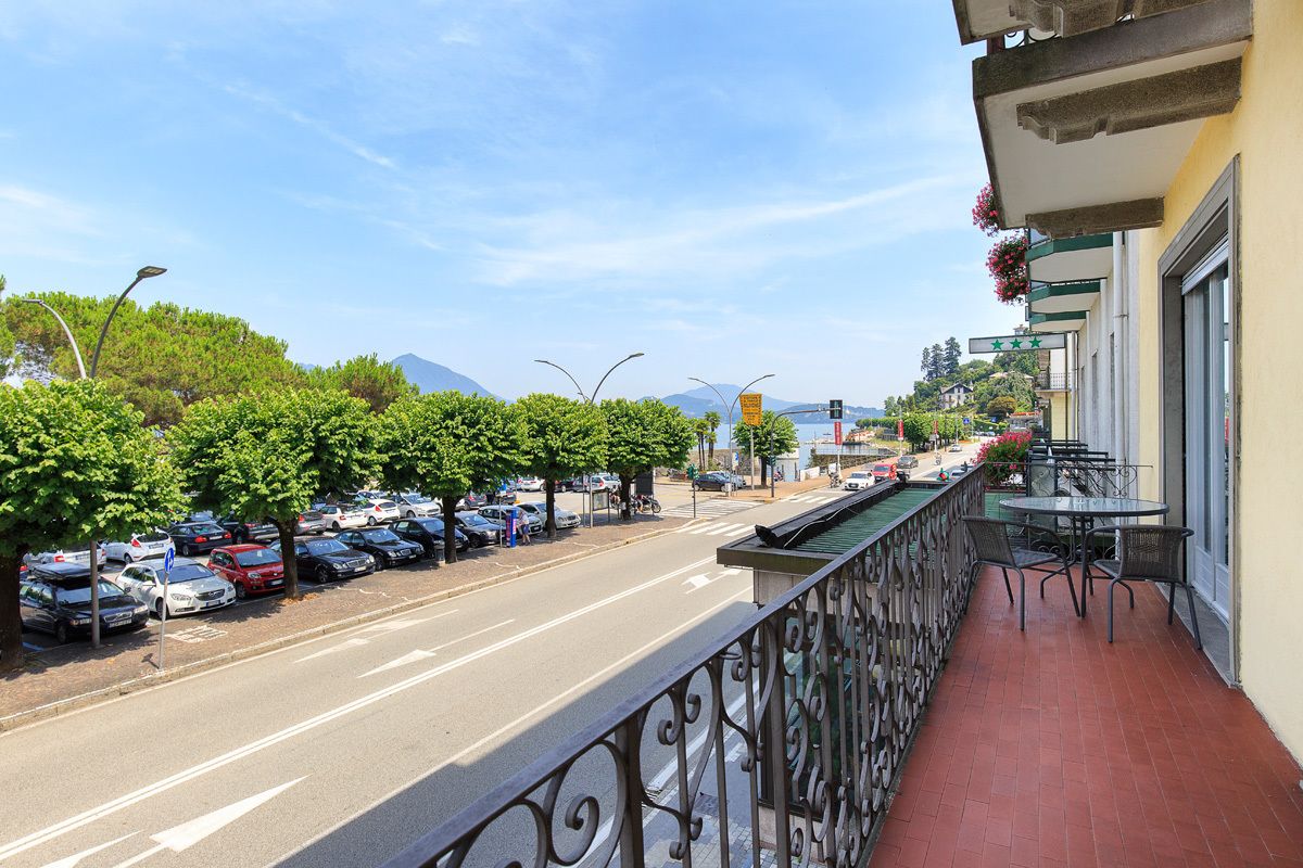 A balcony with a view of a street lined with parked cars and trees, a restaurant with outdoor seating, and a mountain in the background.