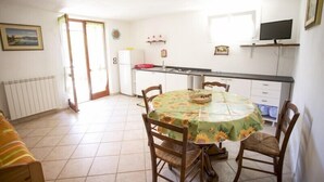 A dining area with a round table covered in a floral tablecloth, surrounded by wooden chairs. A refrigerator and a sink are visible in the background. - Appartamento Il Cannucciato (Portoferraio)