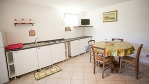 A compact kitchen with white cabinets, a small dining table with chairs, a TV on a shelf, and a window providing natural light. - Appartamento Il Cannucciato (Portoferraio)