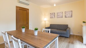 A dining area with a wooden table, white chairs, a grey sofa, and a wall-mounted air conditioner. - Impero House Rent - Adelaide (Stresa)