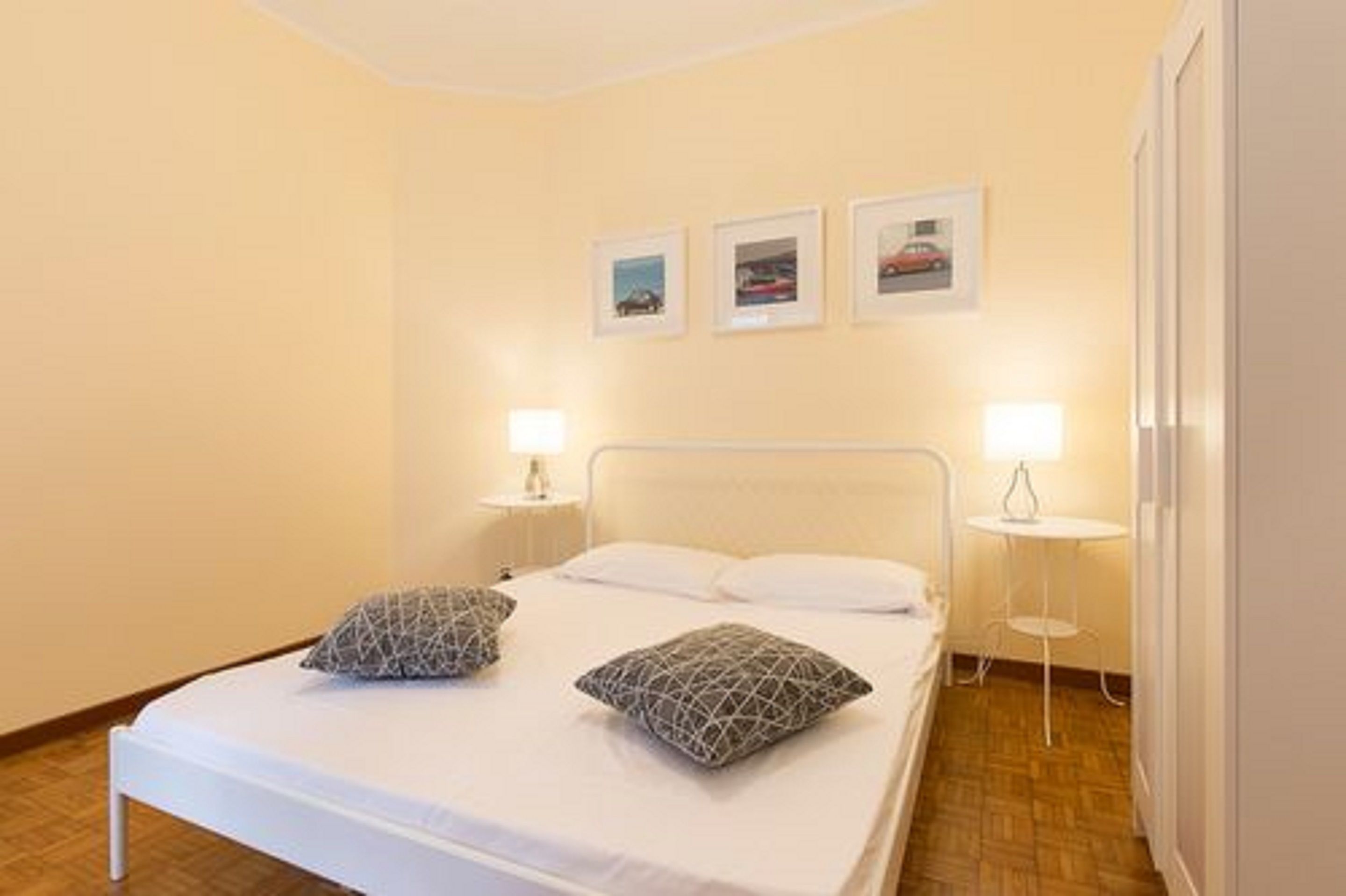 Ein ordentlich bezogenes Bett mit zwei Kissen, ein Nachttisch mit Lampe und drei gerahmte Bilder an der Wand.