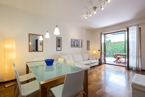 A modern living room with a glass dining table, white sofas, and a view of a garden through large sliding doors. - Impero House Rent - Monte Grappa (Stresa)