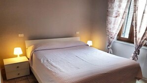 A neatly made bed with a light-colored bedspread, two bedside lamps, and a window with curtains. - Appartamento Bilo San Rocco (Portoferraio)