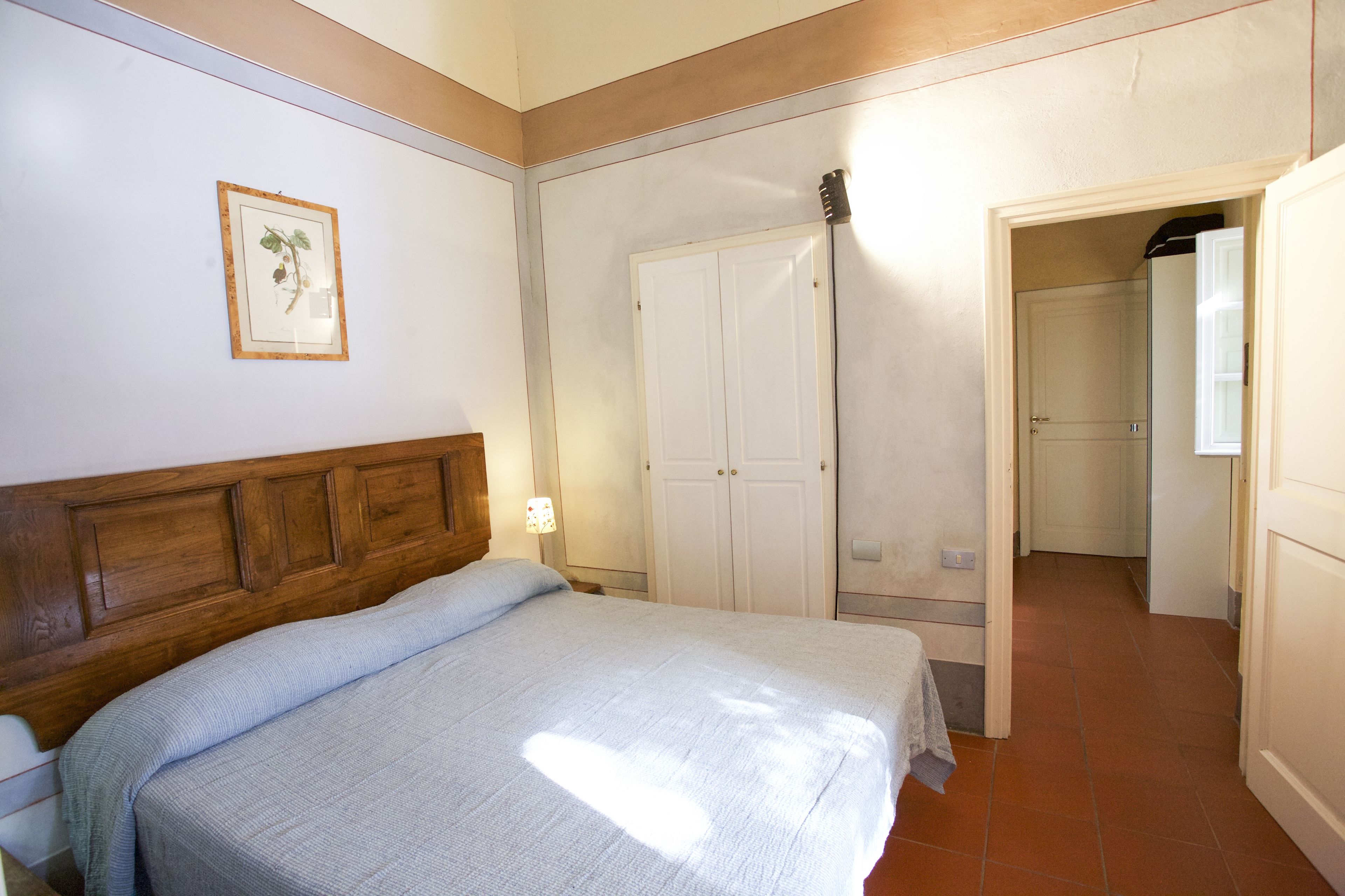 A bedroom with a wooden headboard, a bed with a white bedspread, a framed botanical print on the wall, and a white wardrobe.