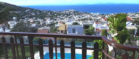 A balcony with a view of a coastal town, a swimming pool, and a mountain range in the distance.