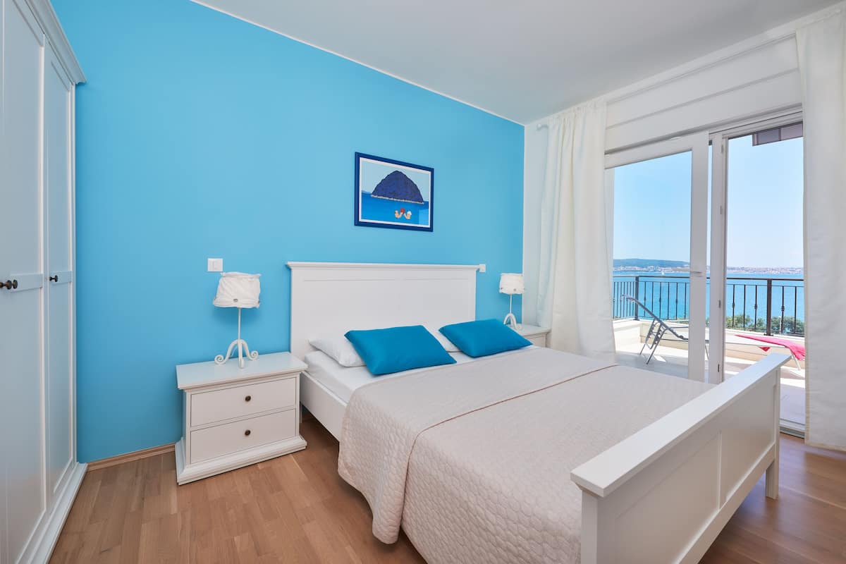 A bedroom with a bed, a nightstand, a window with a view of the sea, and a picture on the wall.