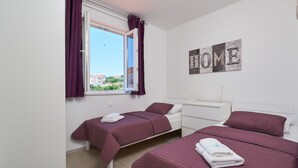 Habitación de hotel con dos camas, una ventana con cortinas moradas, un tocador blanco y un letrero en la pared que dice 'HOME'.