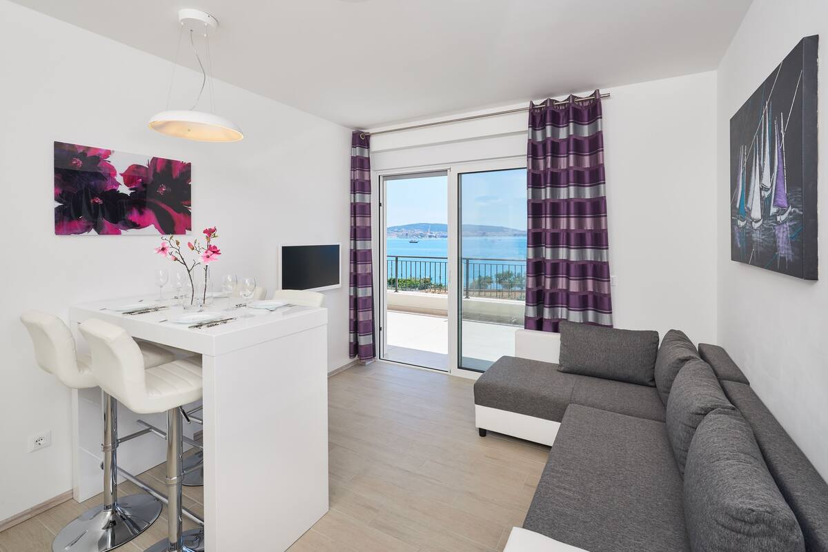 A modern living room with a grey sofa, a white dining area with bar stools, a flat-screen TV, and a balcony with a view of the sea.