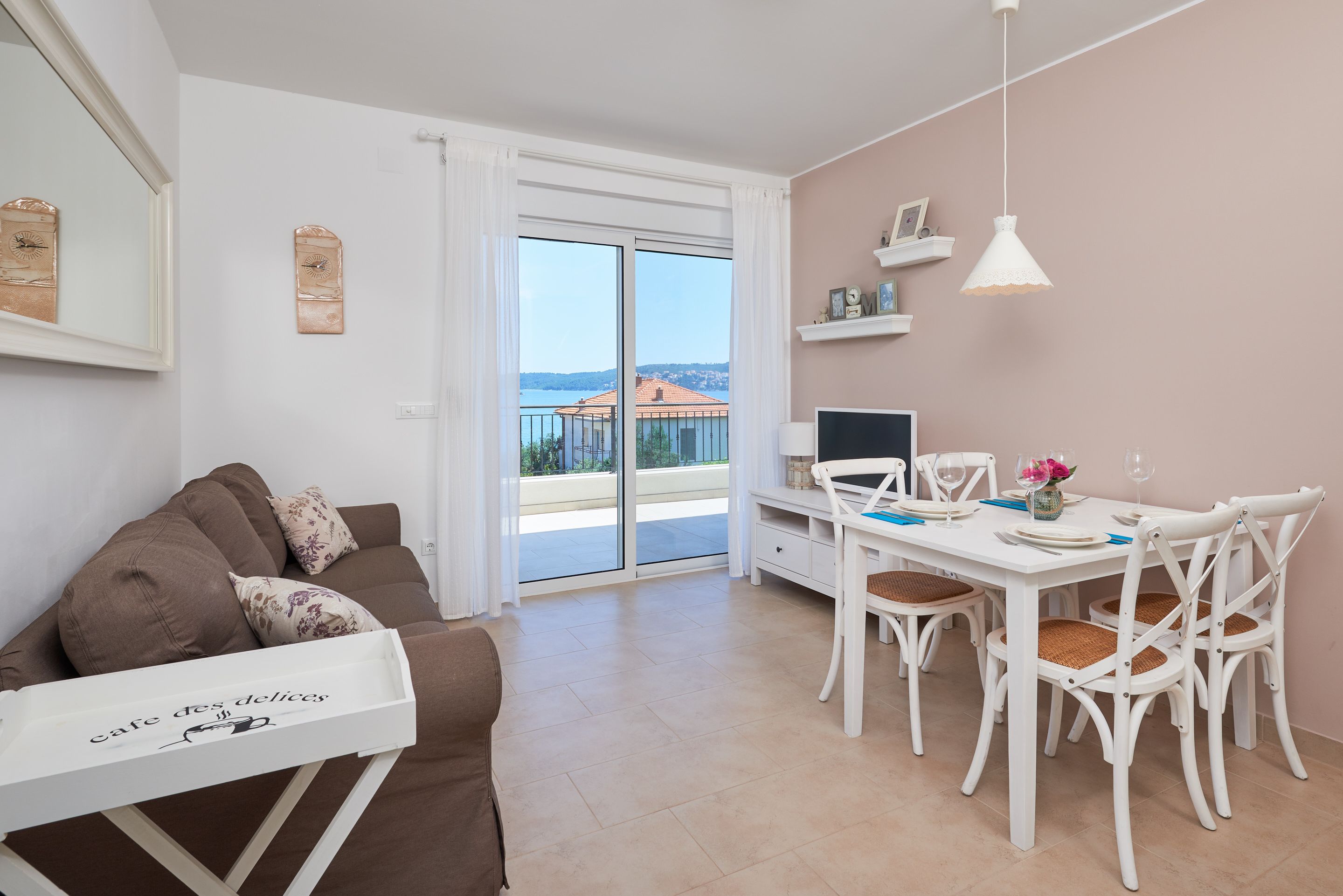 Un salon moderne avec un canapé marron, une table à manger blanche et des chaises. On y trouve un téléviseur posé sur un meuble blanc, un luminaire suspendu blanc et une vue sur la mer par de grandes fenêtres.