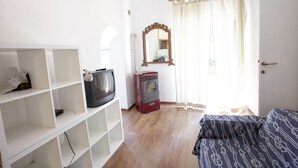 Une pièce avec un plancher en bois, un lit recouvert d’une literie à motifs, un téléviseur posé sur une étagère blanche, un radiateur rouge, un miroir et une porte avec un rideau.