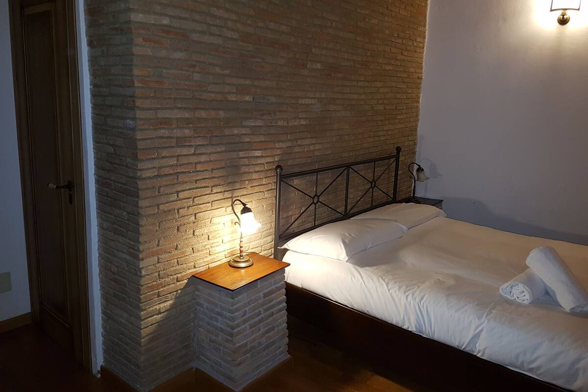 Ein Schlafzimmer mit Backsteinwand, einem Holznachttisch mit Lampe, einem Bett mit weißer Bettwäsche und einem Handtuch auf dem Bett.