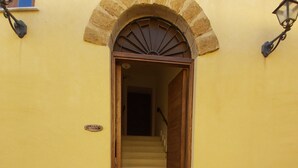 Uma escadaria leva a uma porta com arco de pedra, um pequeno letreiro na parede e duas luminárias fixadas na parede.