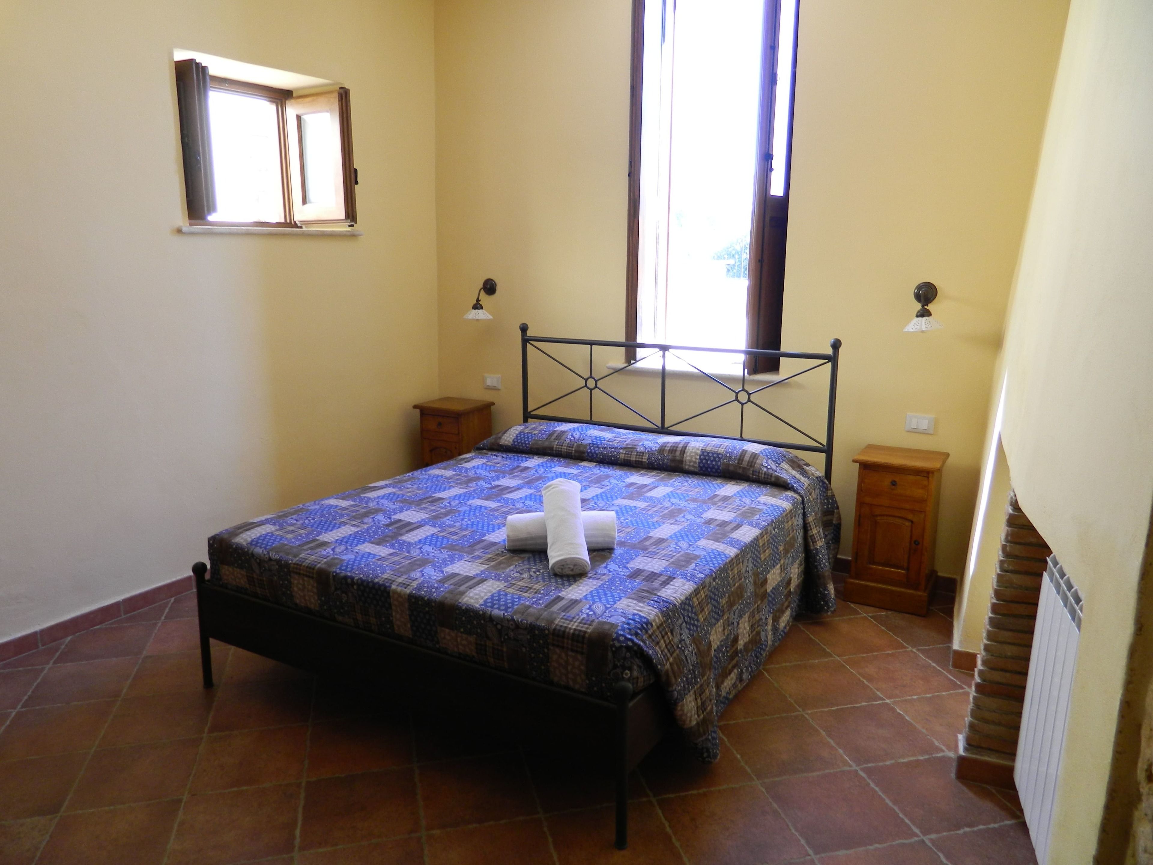 Un dormitorio con cama, ventana, mesita de noche y una lámpara de pared.