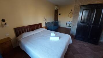 Una cama bien hecha con una colcha blanca y dos toallas, un cabecero de madera, una mesita de noche, una lámpara de pared, una pequeña mesa con una silla, un lavabo y una puerta de madera oscura.