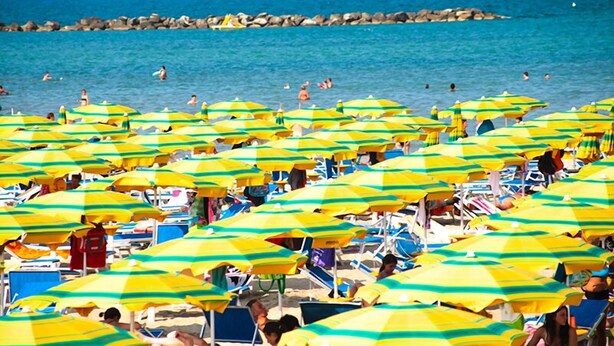 A beach with numerous yellow and green striped umbrellas, people sunbathing, and a rocky breakwater in the background.