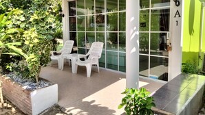 Un patio couvert avec deux chaises en plastique blanc et une table, entouré de plantes en pot et d’une grande porte vitrée donnant sur l’intérieur.