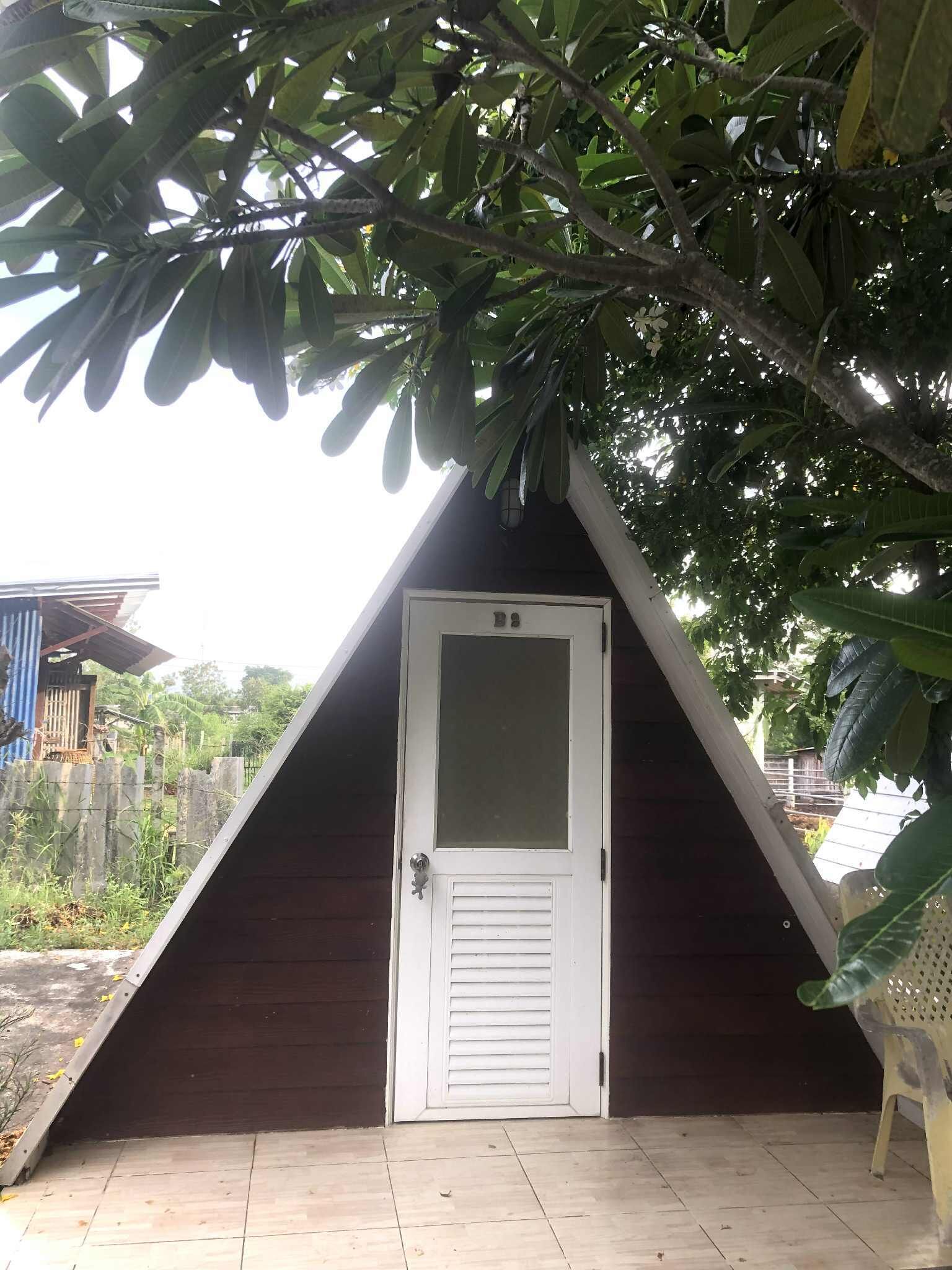 Uma estrutura de madeira triangular com uma porta branca, rodeada por árvores e um chão de azulejos.