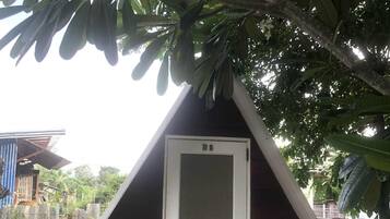 Uma estrutura de madeira triangular com uma porta branca, rodeada por árvores e um chão de azulejos.