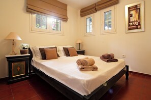 A neatly made bed with a white bedspread and two rolled towels, a bedside table with a lamp and a basket, and a painting on the wall. - Oasis Pool House by GHH (Saronikos)