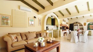A spacious living room with a beige sofa, a wooden coffee table with candles, and a kitchen area with a dining set. - Oasis Pool House by GHH (Saronikos)