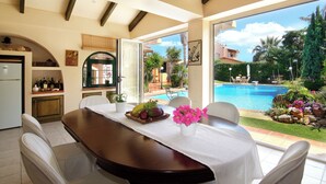 A dining area with a table set for two, a fruit basket, and a vase of flowers. - Oasis Pool House by GHH (Saronikos)