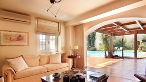 A spacious living room with a beige sofa, a glass coffee table, and a view of a pool area. - Oasis Pool House by GHH (Saronikos)