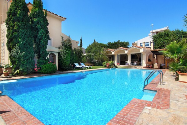 Una piscina con agua azul cristalina, rodeada por un patio con baldosas rojas y un edificio de fachada color beige y ventanas de arco.