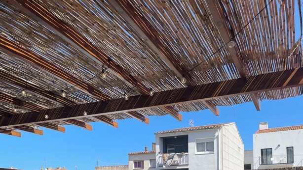 A rooftop patio with a wooden table and chairs, a covered seating area with cushions, and a view of neighboring buildings.