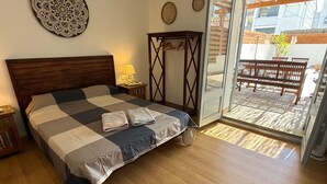 A bedroom with a bed, wooden floor, a wall-mounted decorative item, a small table with a lamp, and a view of the outside balcony.