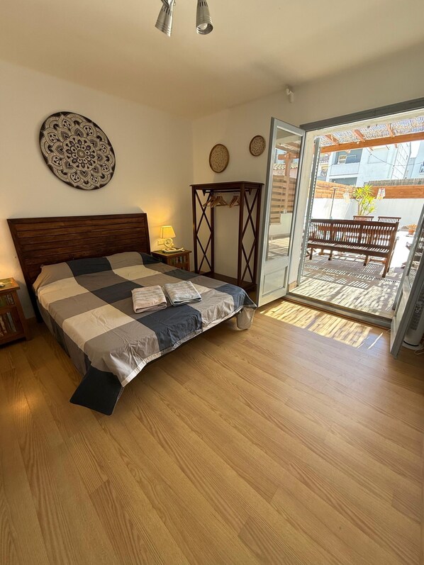 Un dormitorio con cama, suelo de madera, un adorno en la pared, una mesita de noche con lámpara y vistas al balcón exterior.