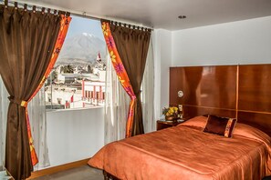 A hotel room with a large bed, a wooden headboard, a nightstand with a vase of flowers, and a view of a mountain and cityscape through sheer curtains.