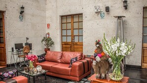 A lobby with a red leather sofa, a glass coffee table, a wooden side table with a vase of flowers, and a large window with a view of the street. - Hotel Tikary (Arequipa)