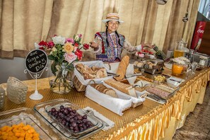 A buffet table with various food items, a sign that reads "Que tenga un buen día," and a person in traditional attire.