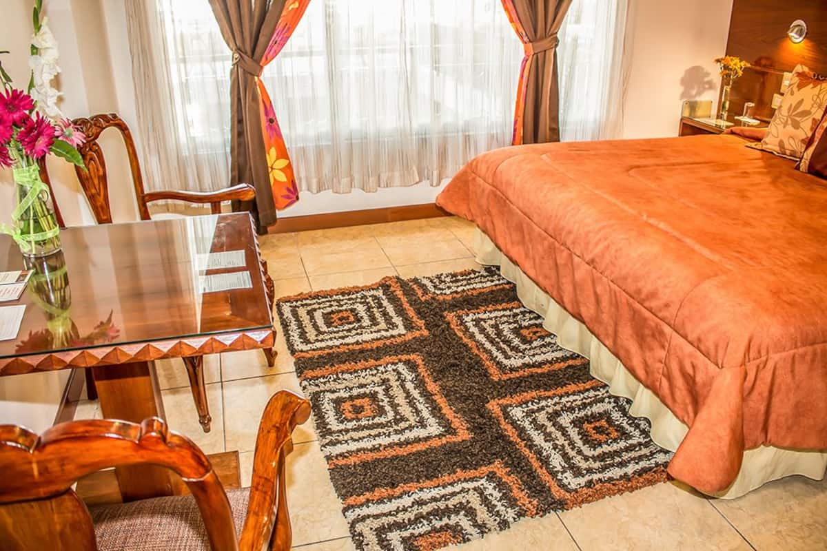 A bedroom with a large bed, a wooden table with chairs, a glass-top table, and a patterned rug.