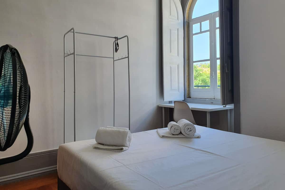 A hotel room with a bed, a window, a chair, and a towel rack.