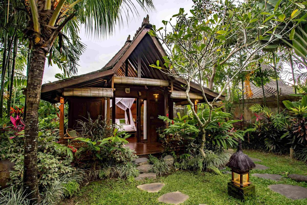 A traditional wooden house with a thatched roof, surrounded by lush tropical vegetation and a stone pathway leading to the entrance.