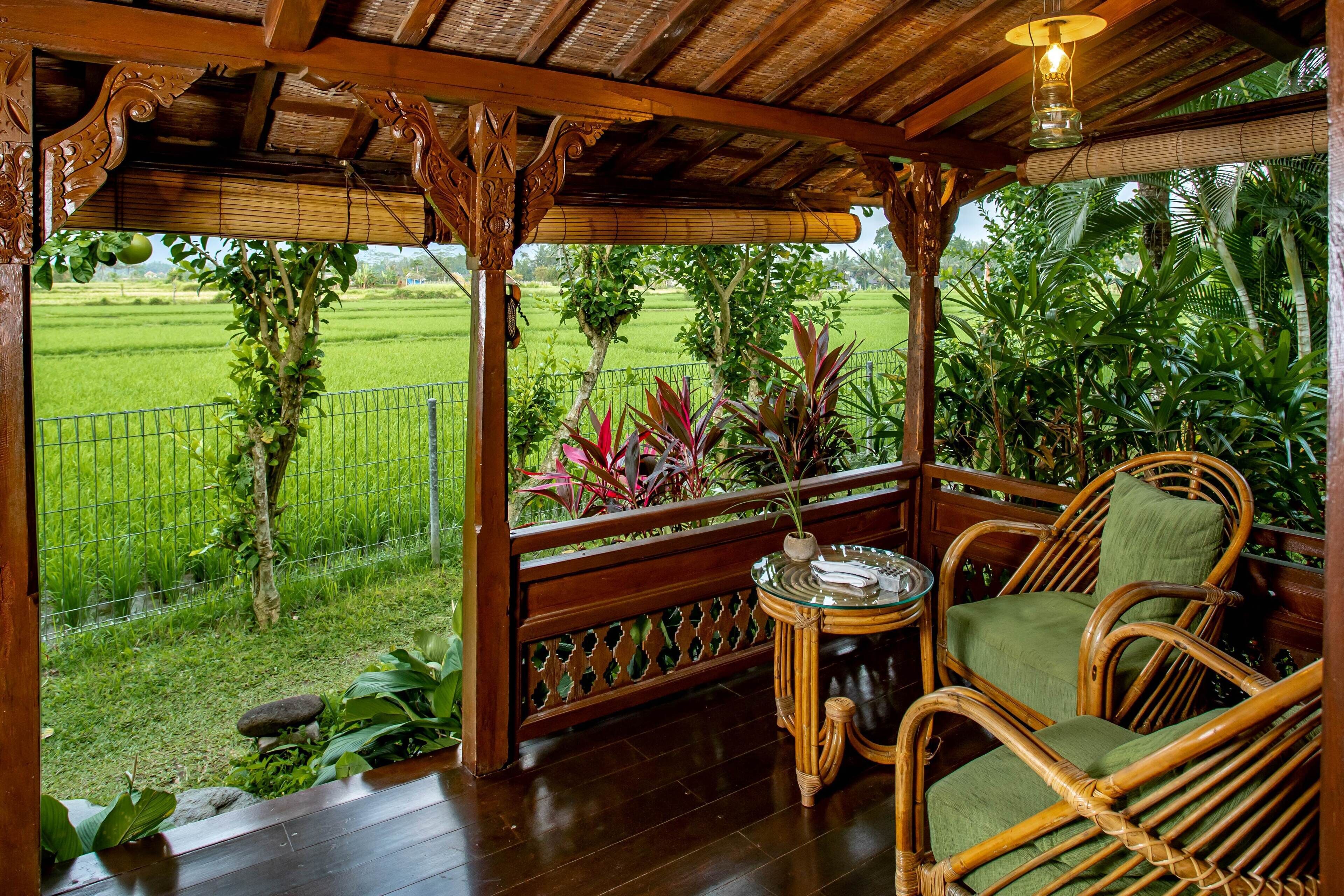 A wooden porch with a glass table and wicker chairs, overlooking a lush garden and a rice field.