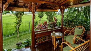 A wooden porch with a glass table and wicker chairs, overlooking a lush garden and a rice field.