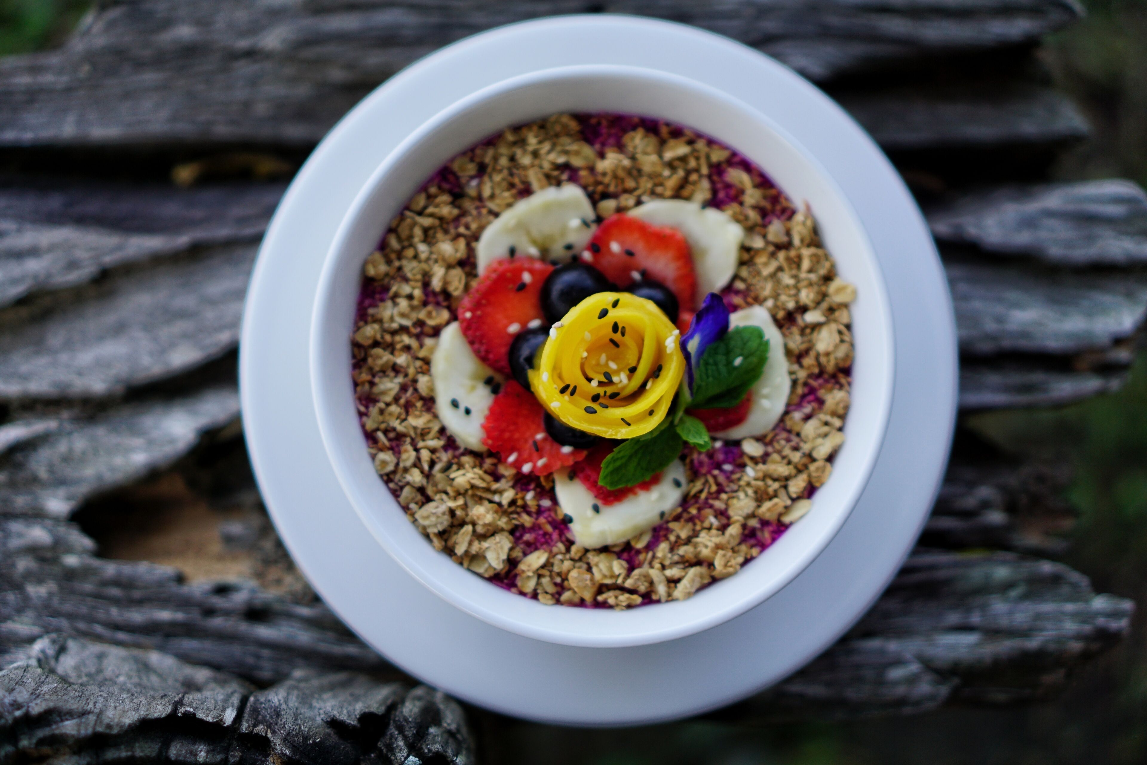 A bowl of granola with fruits and a flower on a wooden surface.