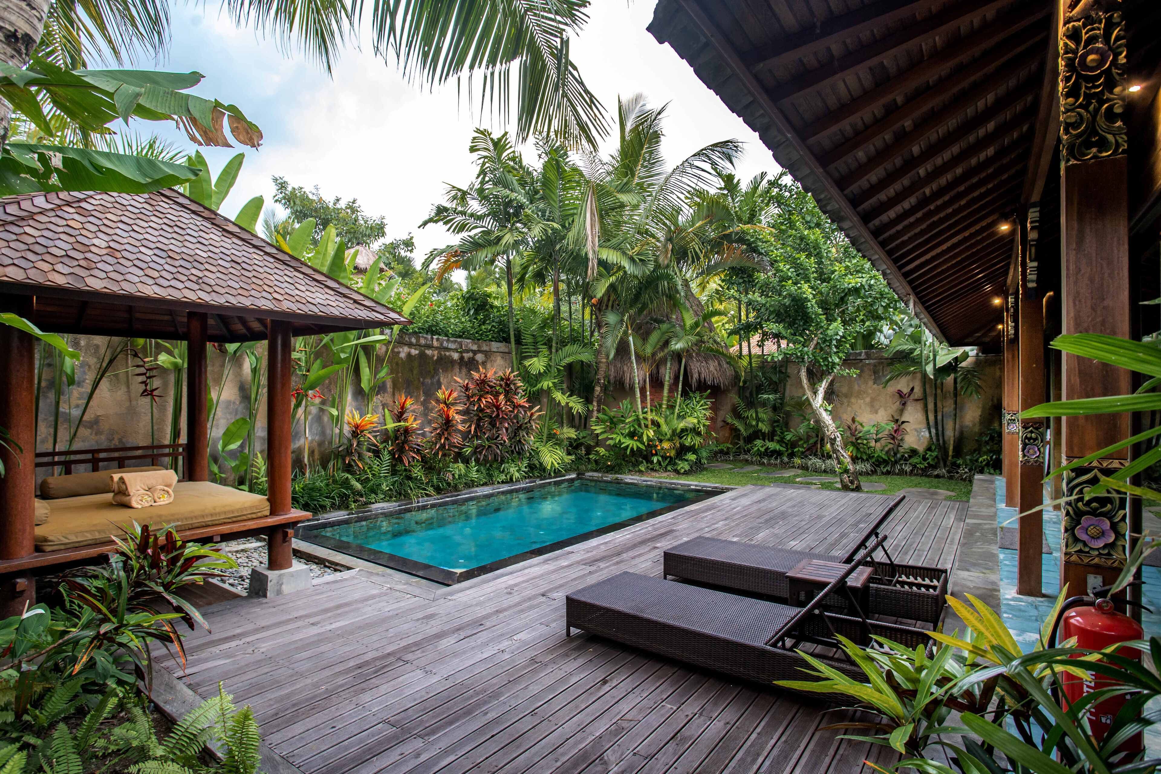 A wooden deck with a pool, lounge chairs, and a covered seating area surrounded by tropical plants.