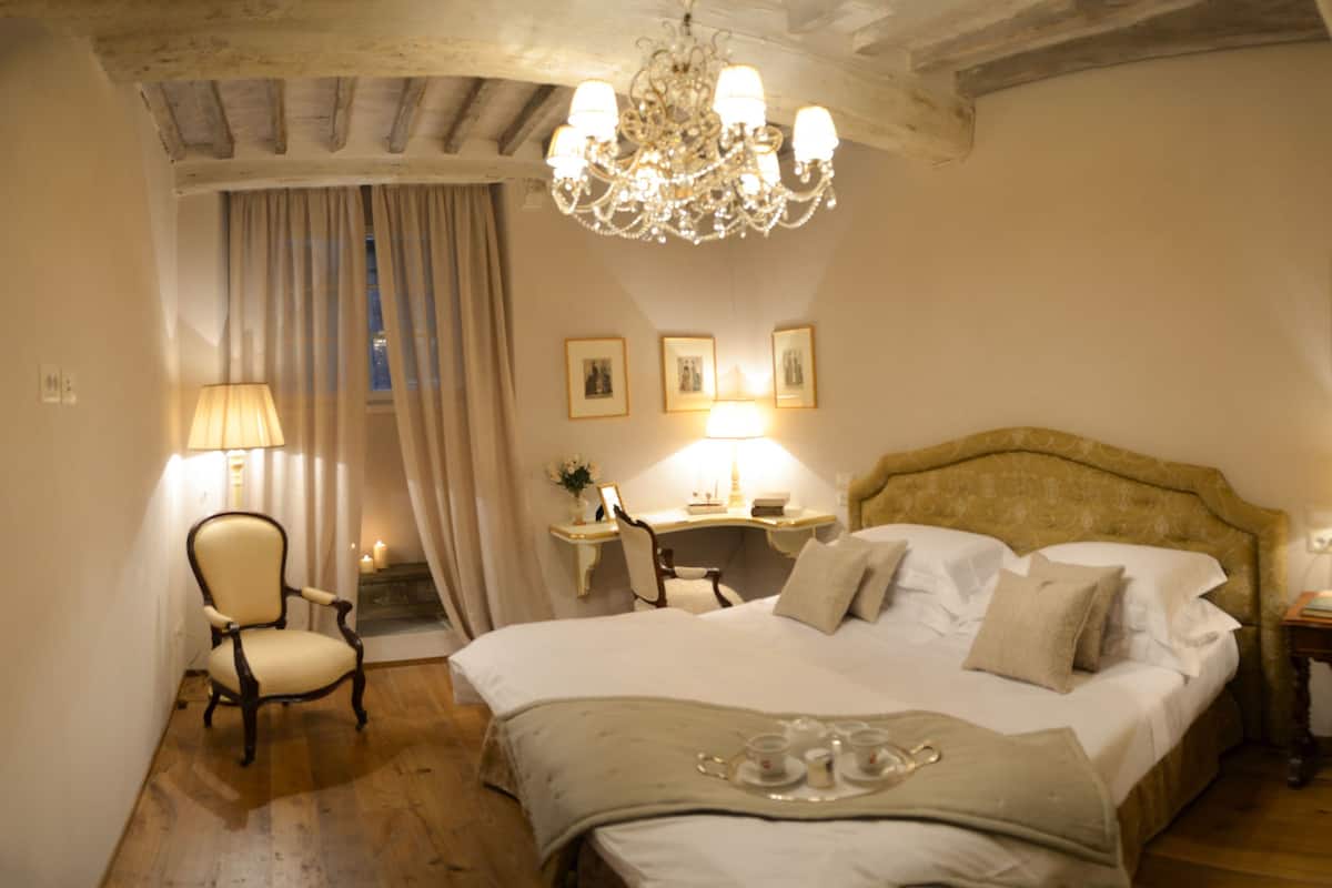 A hotel room with a large bed, a chandelier, a desk with a lamp, and framed pictures on the walls.