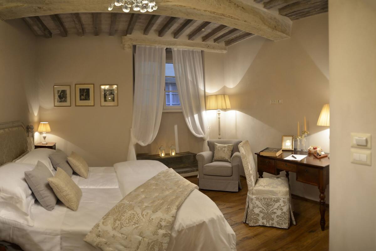 A bedroom with a large bed, two bedside tables with lamps, a chair, a desk, and a window with curtains.
