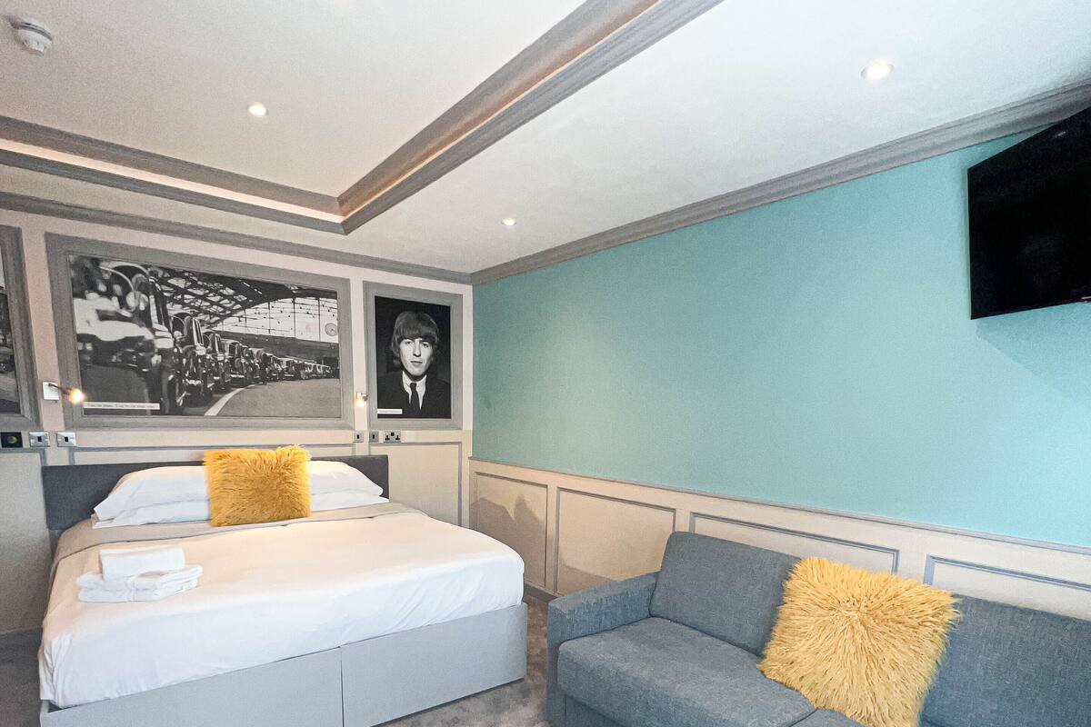 A bedroom with a bed, a grey sofa, a TV mounted on the wall, and a ceiling with recessed lighting.