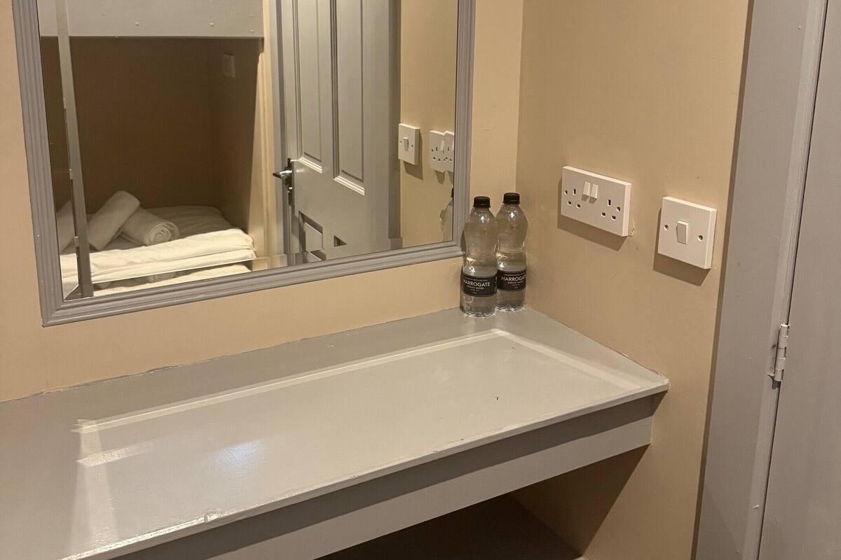 A bathroom with a white sink, mirror, and a bunk bed visible in the reflection. There are two bottles on the sink's edge.