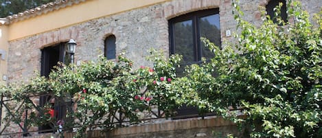 A building with stone walls and a yellow facade, featuring three windows and a small balcony with climbing plants.