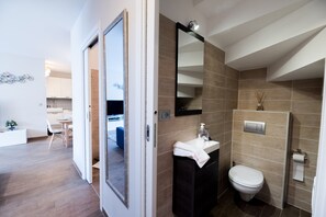 A modern bathroom with a toilet, sink, and mirror. There is a door leading to a dining area with a table and chairs.
